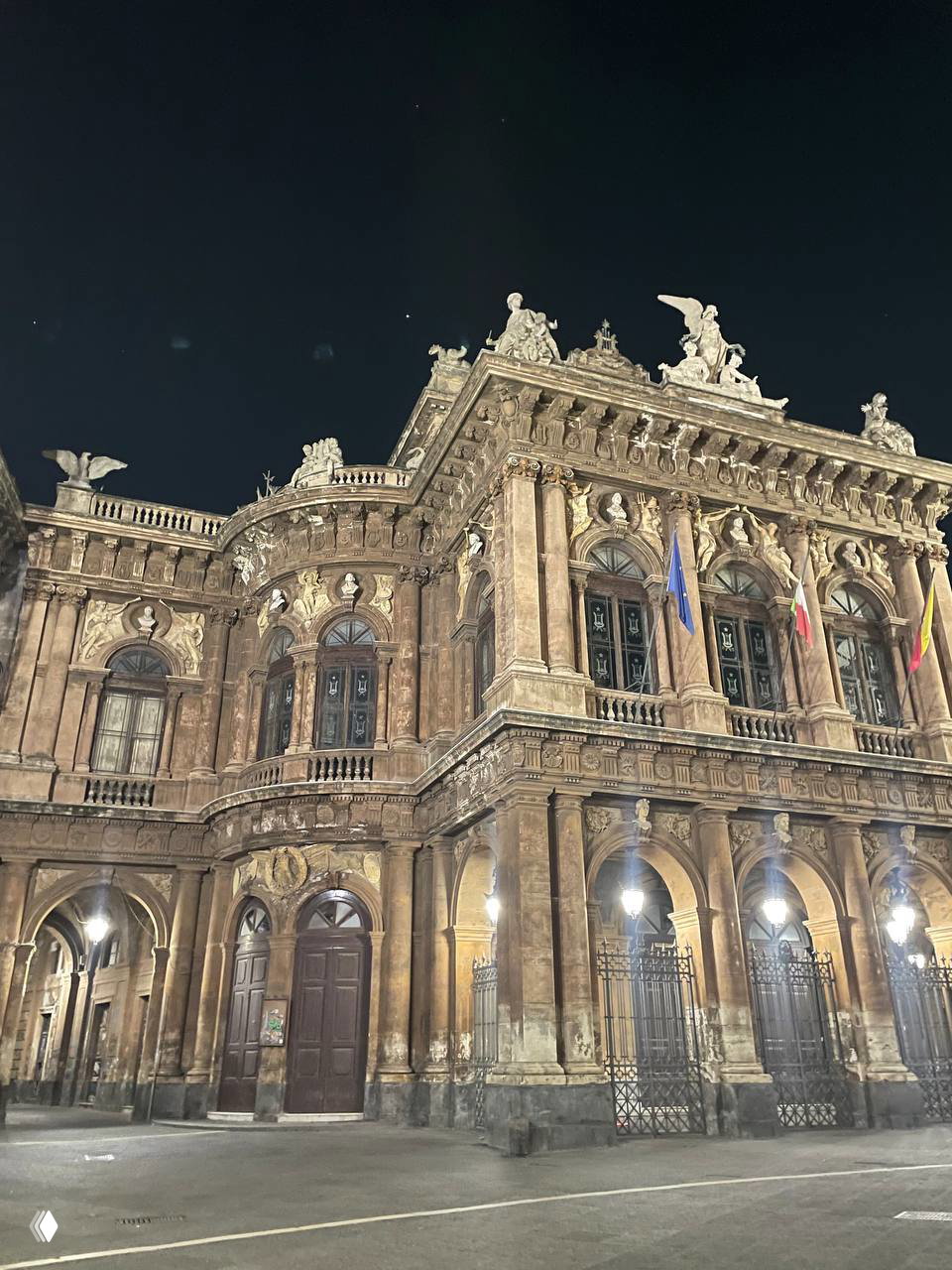 Величественный фасад театрального здания в ночной подсветке, с барельефами, колоннами и вывешенными флагами на площади.