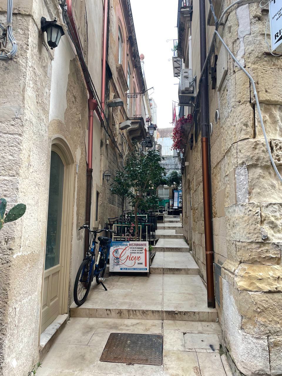 Узкий каменный переулок со ступенями, уличными столиками и припаркованным велосипедом — типичная колоритная улочка Сиракузы.