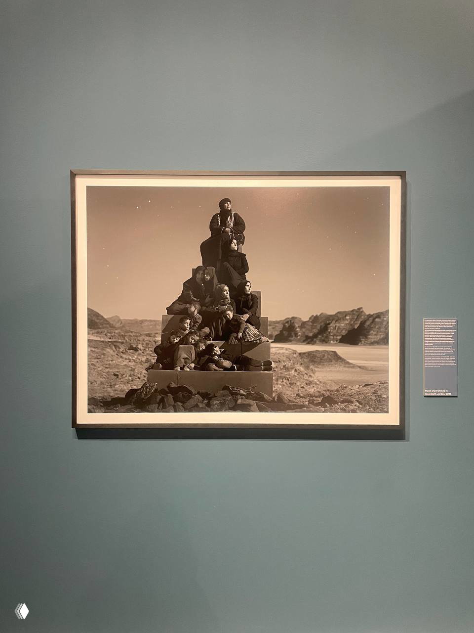 Крупная чёрно‑белая фоторабота с группой людей, сложенных в пирамидальную композицию, висит на голубовато‑зелёной стене Galleria d’Italia.