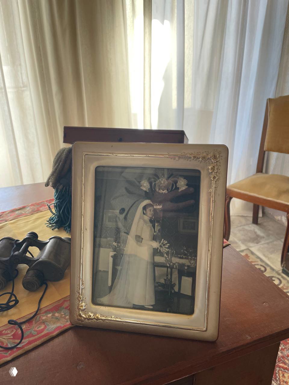Рамка с чёрно‑белой старой фотографией невесты на столе в комнате гостиницы; рядом лежат бинокль и текстильная салфетка — интерьерный объект, связанный с историей дома.