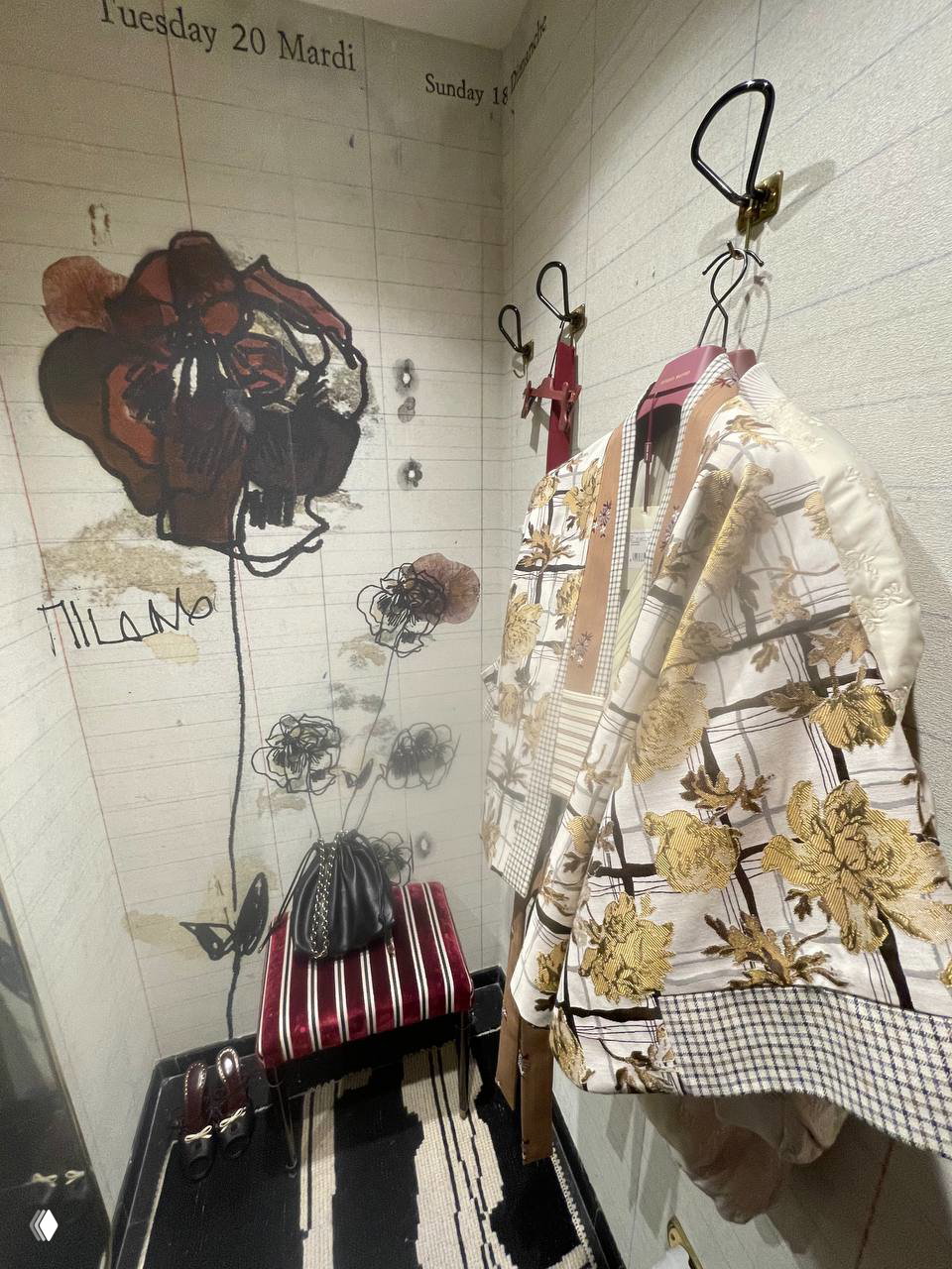 Интерьер примерочной с графическим рисунком на стене и полосатой скамьёй; на вешалке висит цветочный жакет Antonio Marras и сумка на стуле.