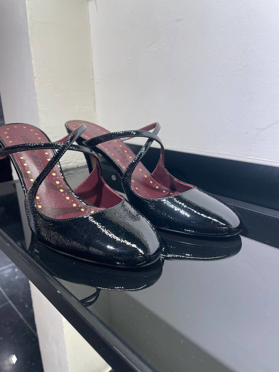 Крупный план блестящих лаковых туфель на каблуке на полке примерочной: чёрные туфли с ремешками и отражением на глянцевой поверхности.