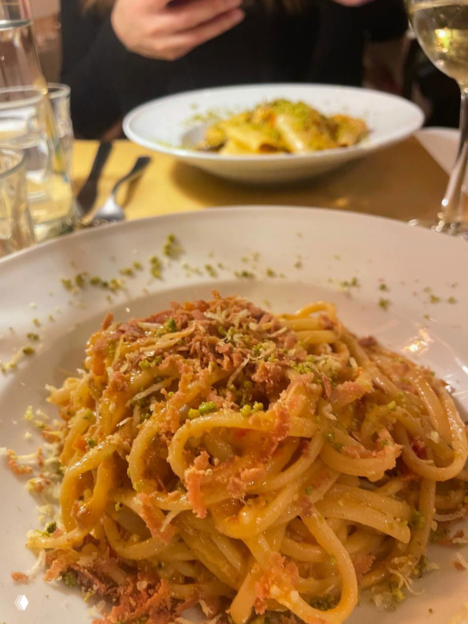 Блюдо пасты на переднем плане с хрустящей посыпкой и фисташками, на заднем плане — ещё одна тарелка и бокал белого вина.