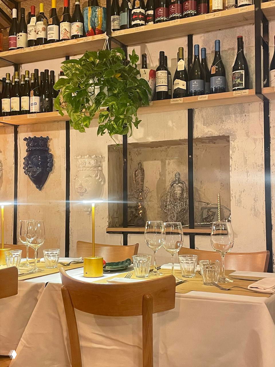 Уютный зал бистро: деревянные стулья, сервированные столы, полки с бутылками вина и висящие декоративные растения над столами.