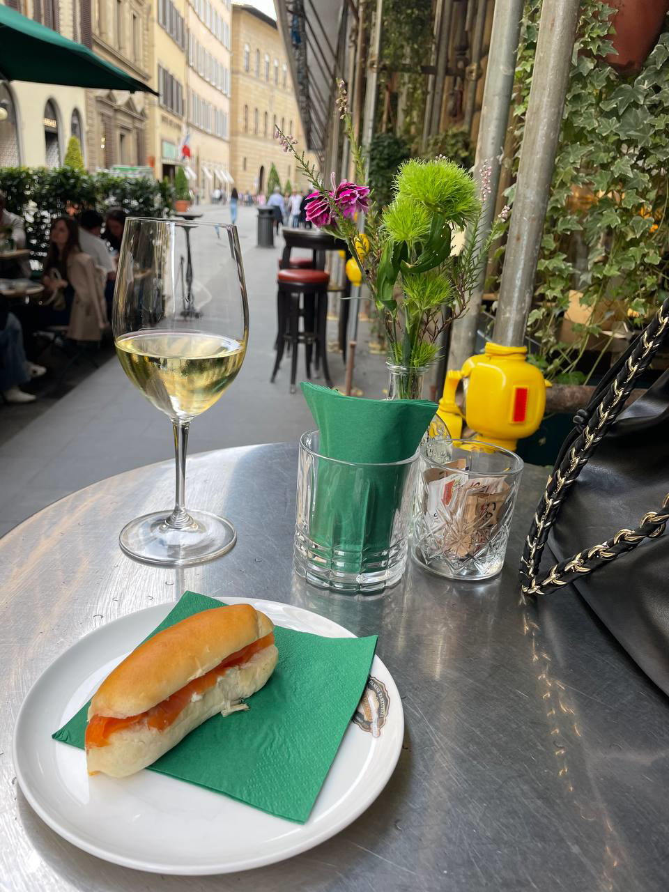 Уличное кафе во Флоренции: бокал белого вина, мини-бутерброд на тарелке, зелёные салфетки и вазочка с цветами на фоне городской улицы и прохожих.