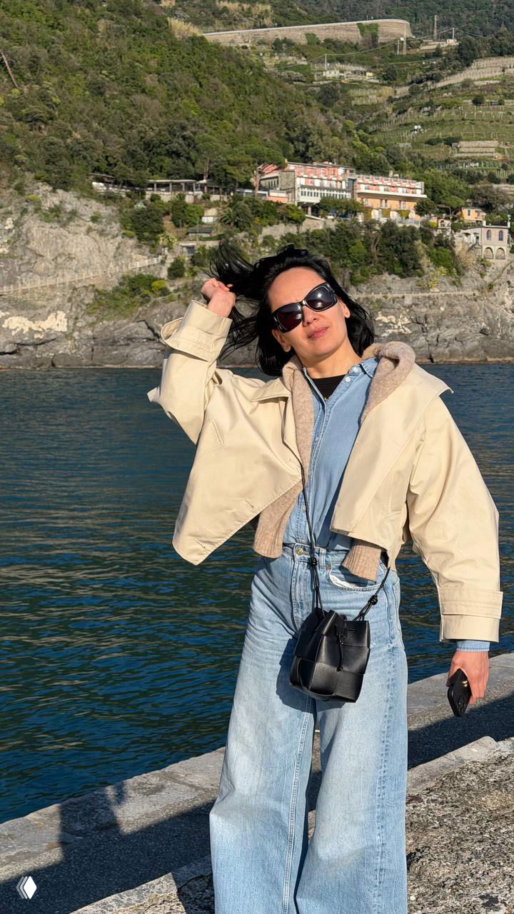 Портрет женщины в светлой куртке и солнцезащитных очках на набережной Cinque Terre на фоне моря, склонов и домиков.