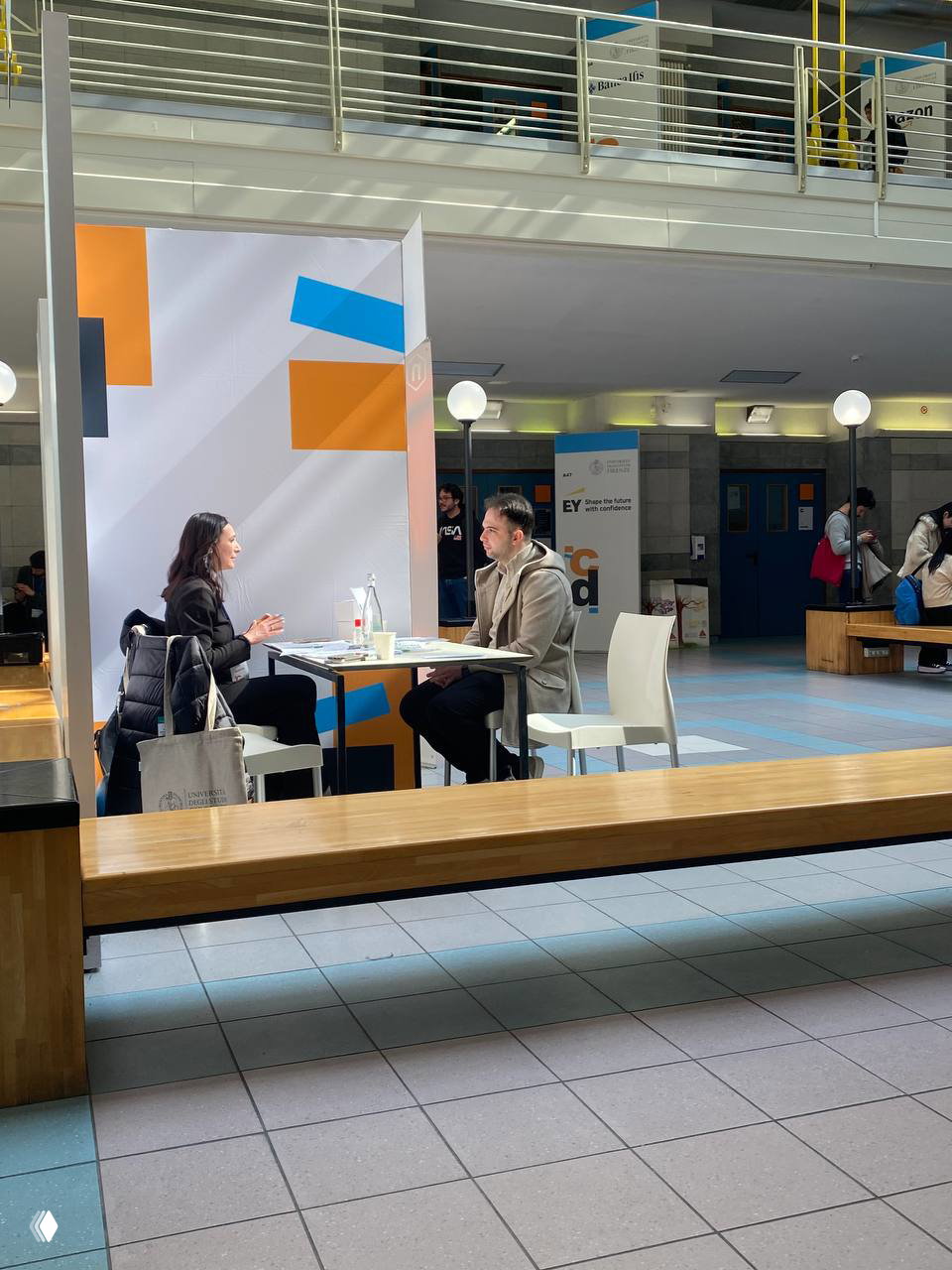 Два человека сидят за столом на карьерном мероприятии: интервью или консультация, рядом информационный стенд и белые стулья в корпусе университета.