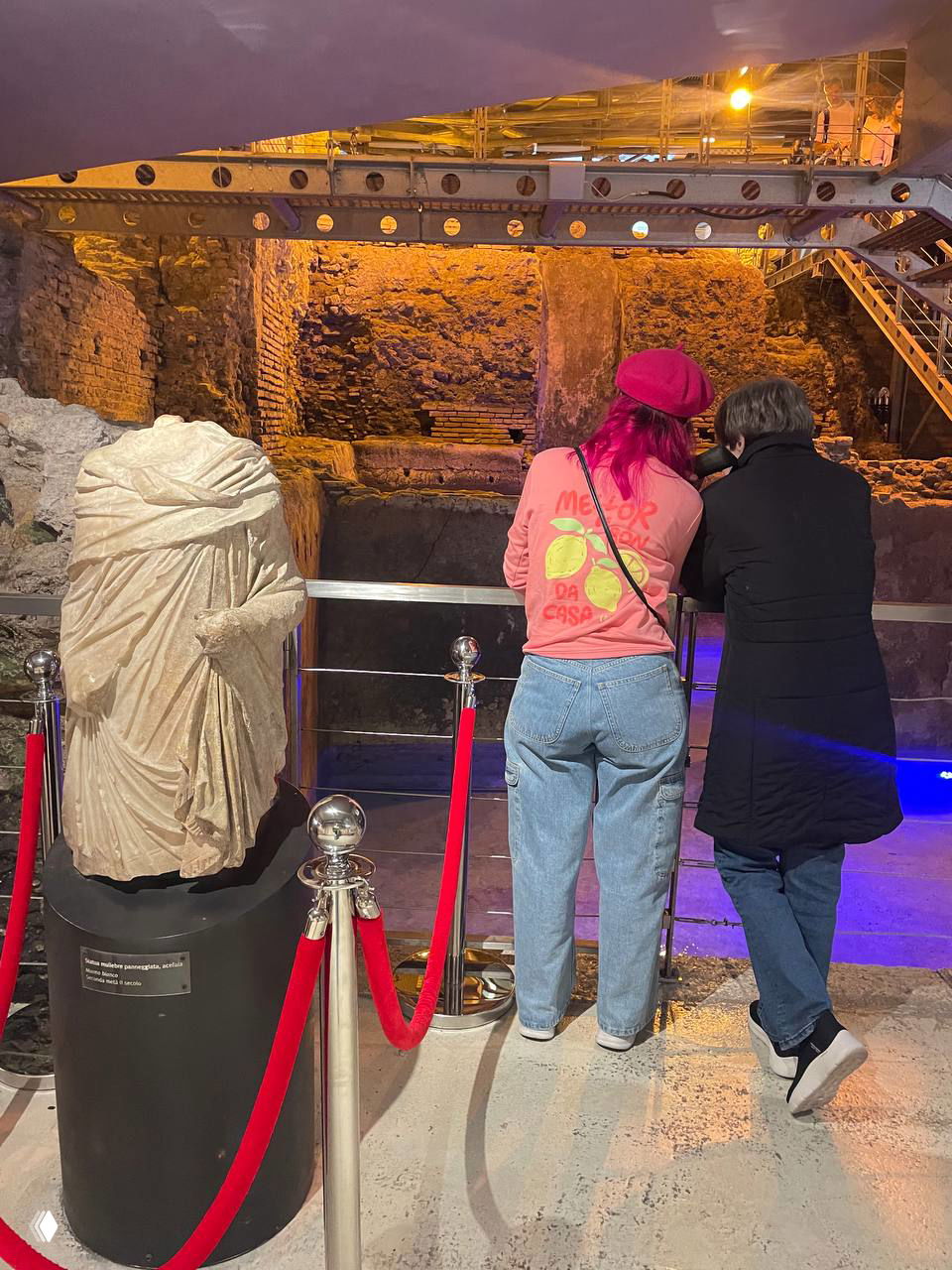 Посетители у ограждения в музее под фонтаном: девушка в розовой толстовке и сопровождающий смотрят на руины и статую на фоне подсвеченных остатков домус.