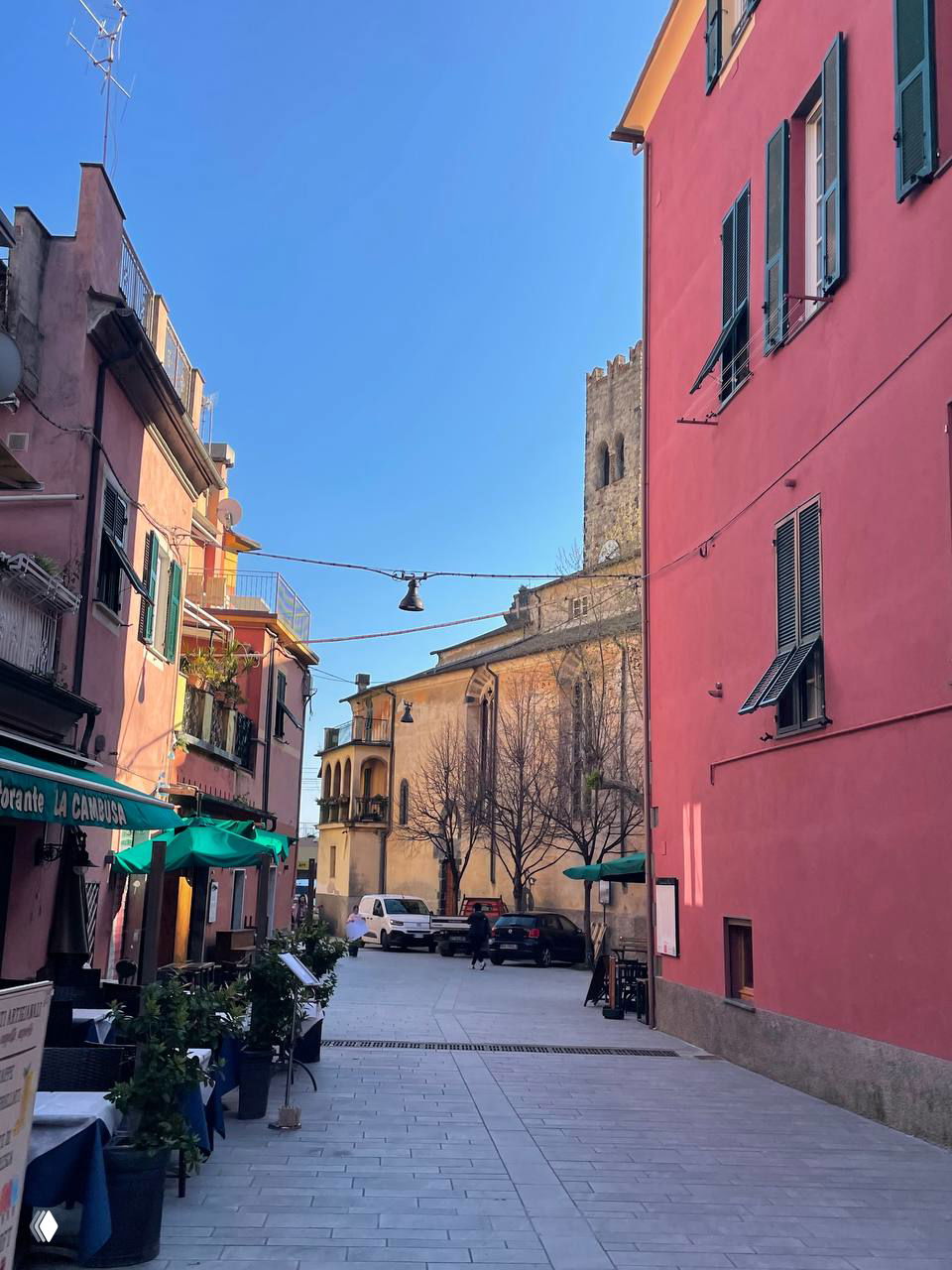 Узкая пешеходная улица в Cinque Terre с розовыми домами, столиками кафе и аккуратно мощёной площадью под ясным голубым небом.