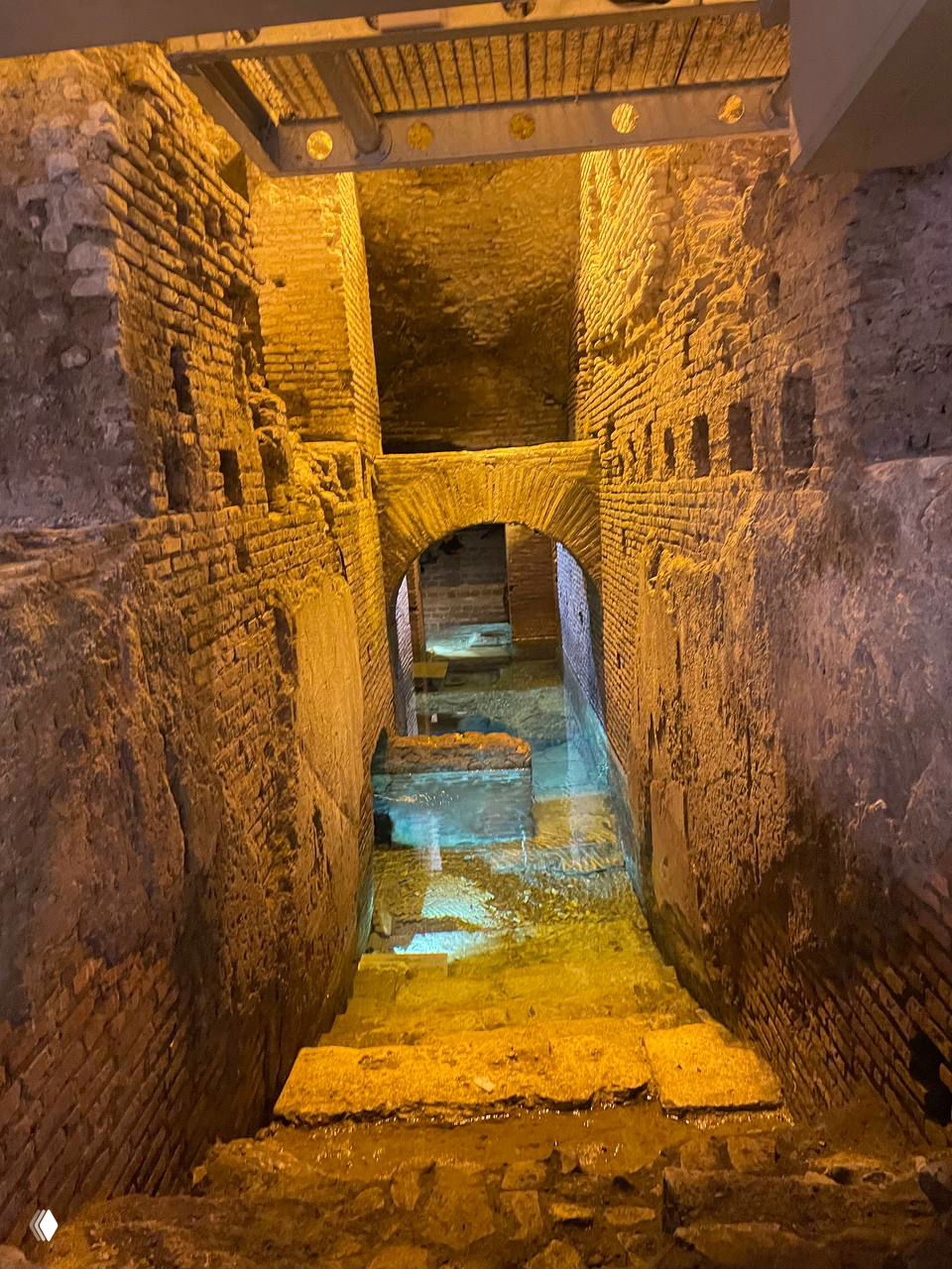 Подсвеченный каменный проход и лестница внутри акведука Aqua Virgo: арки, кирпичная кладка и следы древнего водного канала с остатками воды.