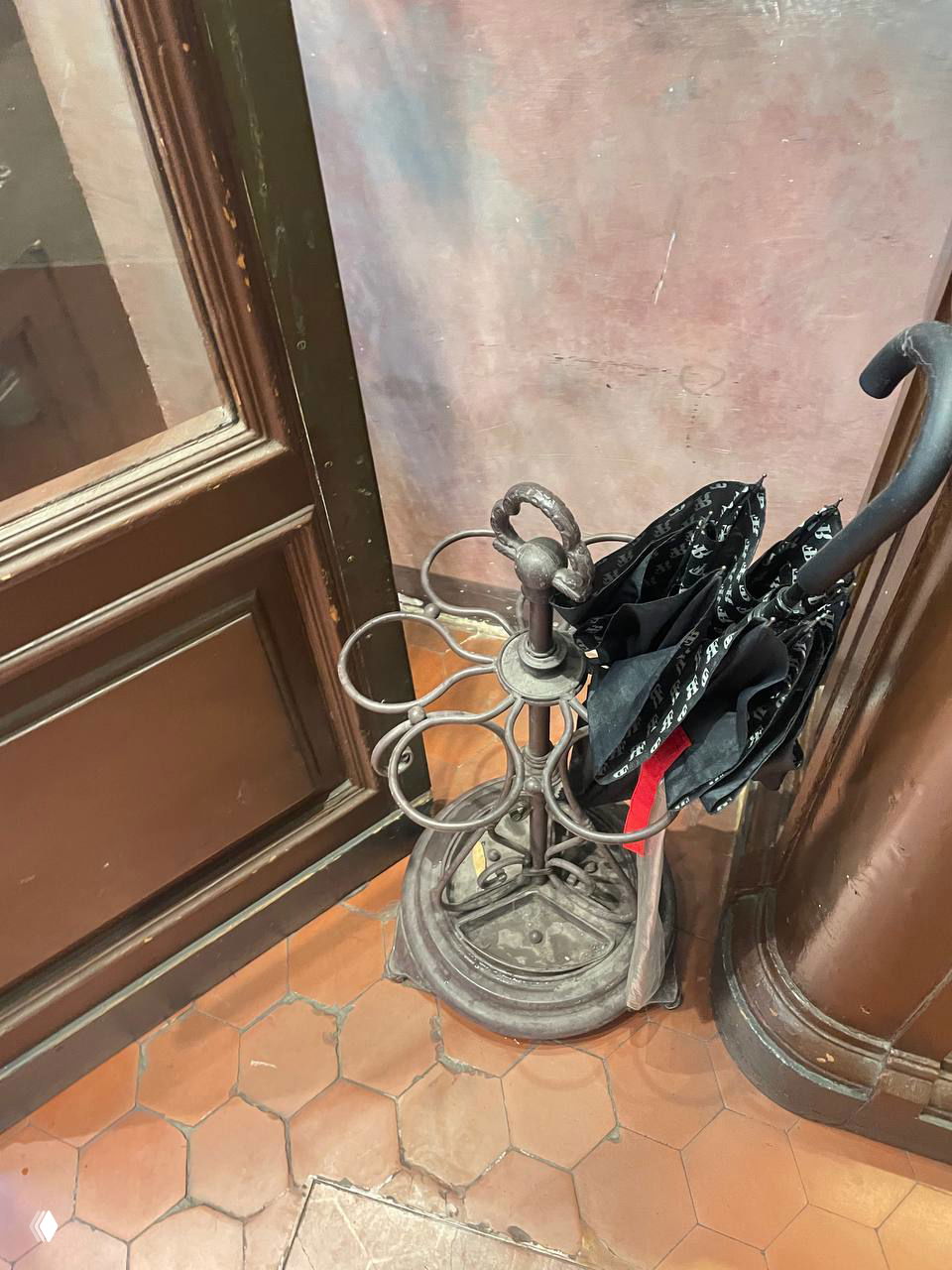 Антикварная металлическая подставка для зонтов у входа: несколько сложенных чёрных зонтов на терракотовой плитке рядом с деревянной дверью.