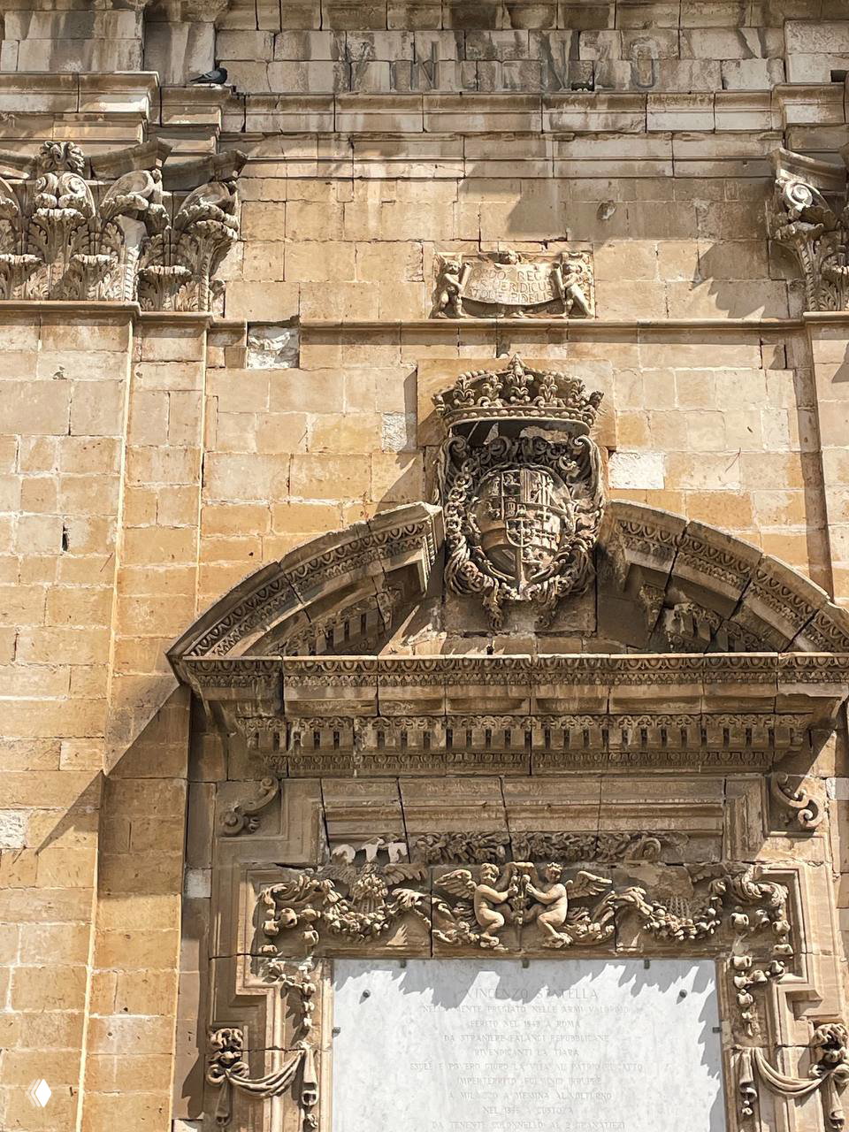 Каменная архитектурная деталь с гербом, резьбой и орнаментом на фасаде исторического здания под дневным светом.