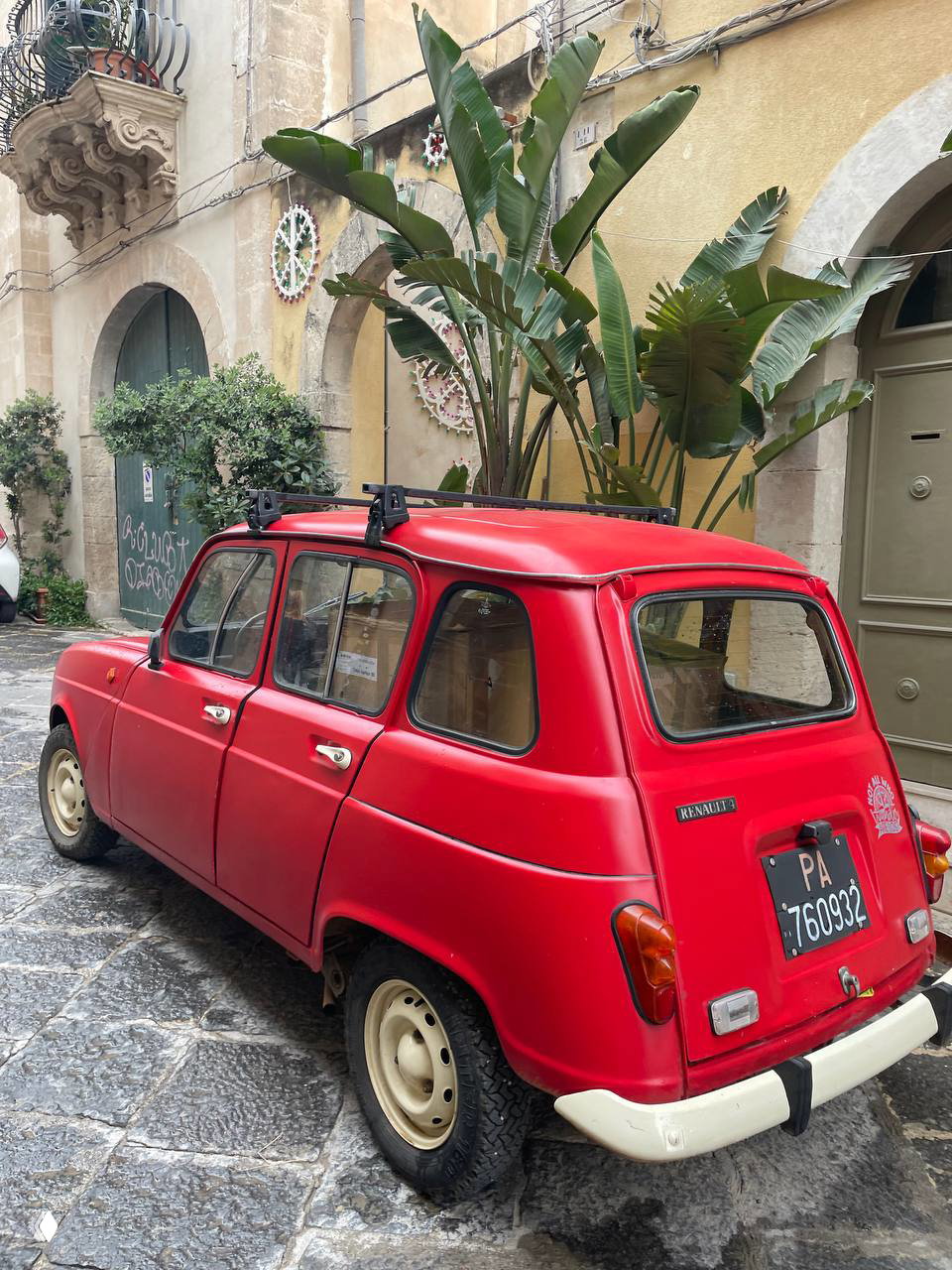 Красный ретро-автомобиль, припаркованный на брусчатке у старых домов и тропических растений — фотокадр сицилийской улицы.