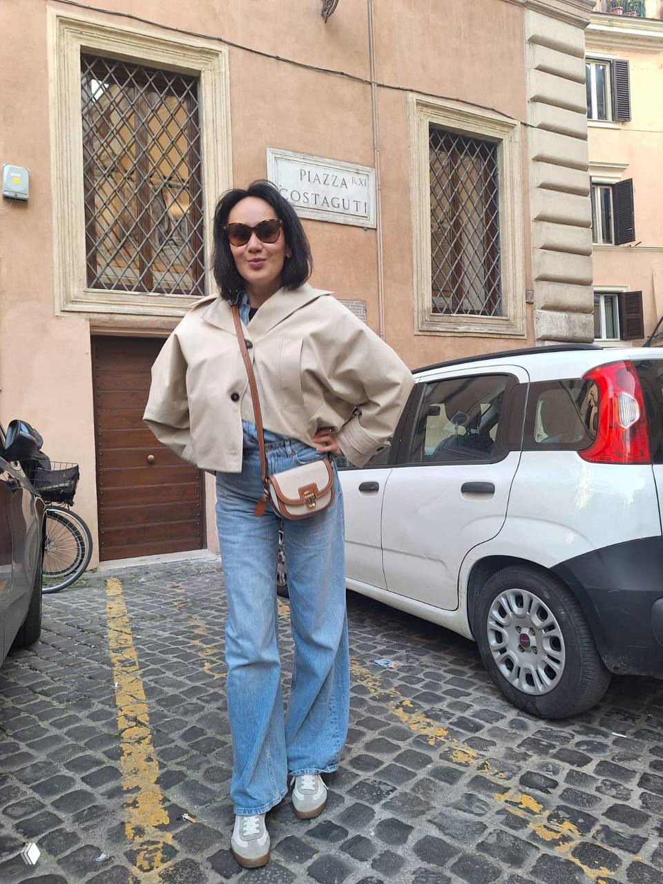 Человек позирует на мощёной площади возле таблички Plaza Costaguti — место съёмок сериала, у здания с типичной римской архитектурой на заднем плане.