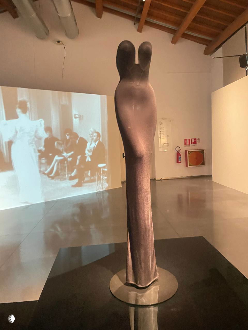 Струнный тёмный манекен с удлинённым облегающим платьем на круглой подставке, отражение на чёрном глянцевом подиуме и проекция сзади.