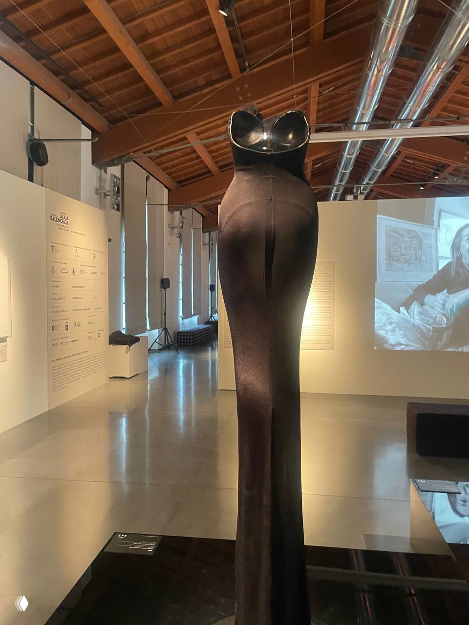 Вид со спины на высокую скульптурную форму платья на манекене: чёткие линии талии и длинный ниспадающий низ, потолочные вентиляционные каналы видны сверху.