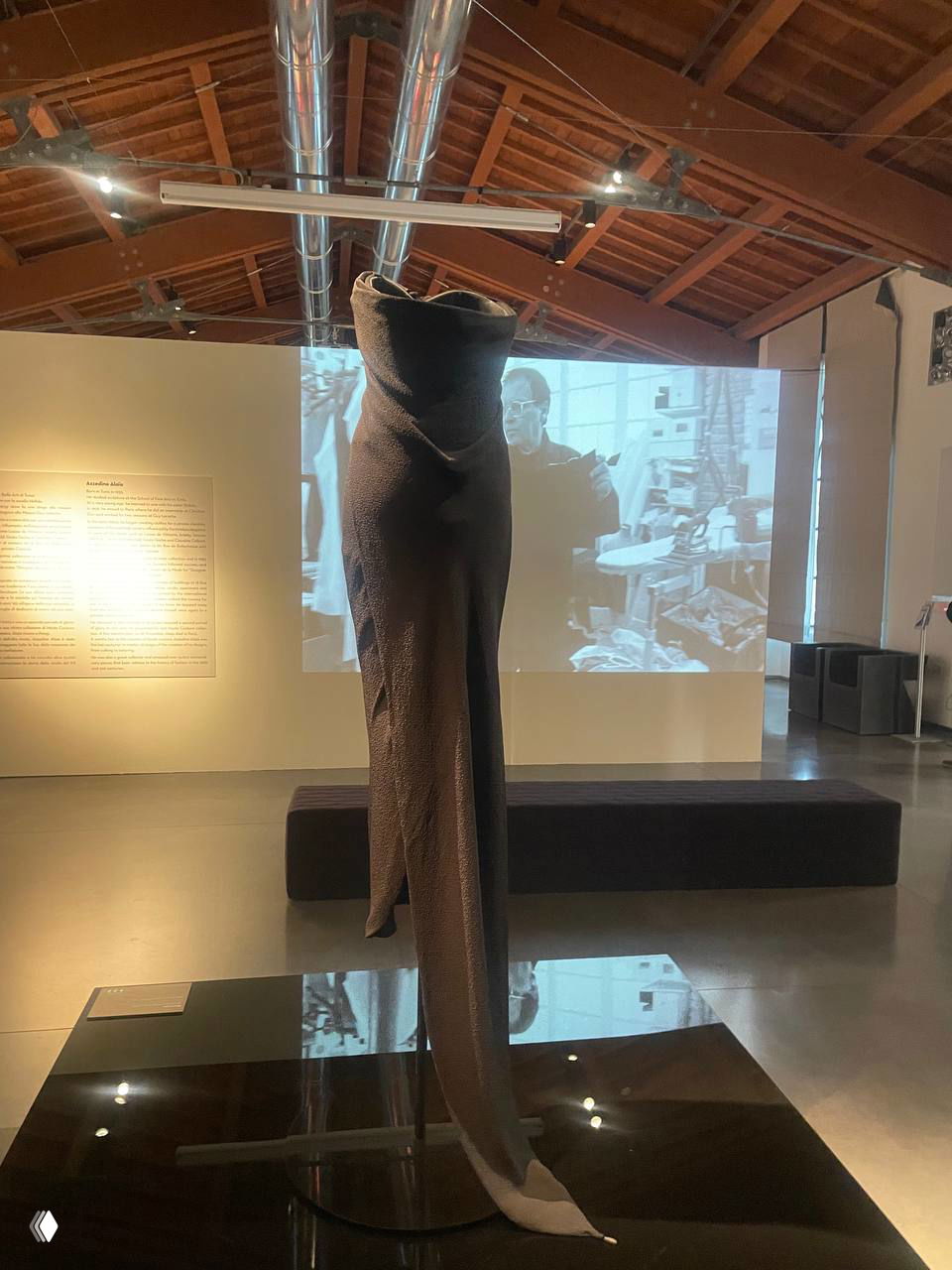 Платье на подиуме, снятое со спины: гладкая чёрная ткань, артистичная посадка, в зале видны окна и плёнки с информацией о выставке.