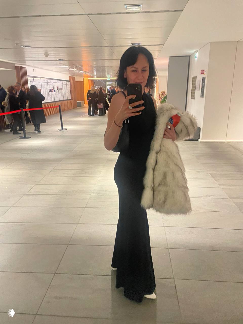 Полнотелое селфи автора в длинном чёрном платье, держащей белую шубу в фойе театра — отражение в зеркале, люди на заднем плане и интерьер фойе.