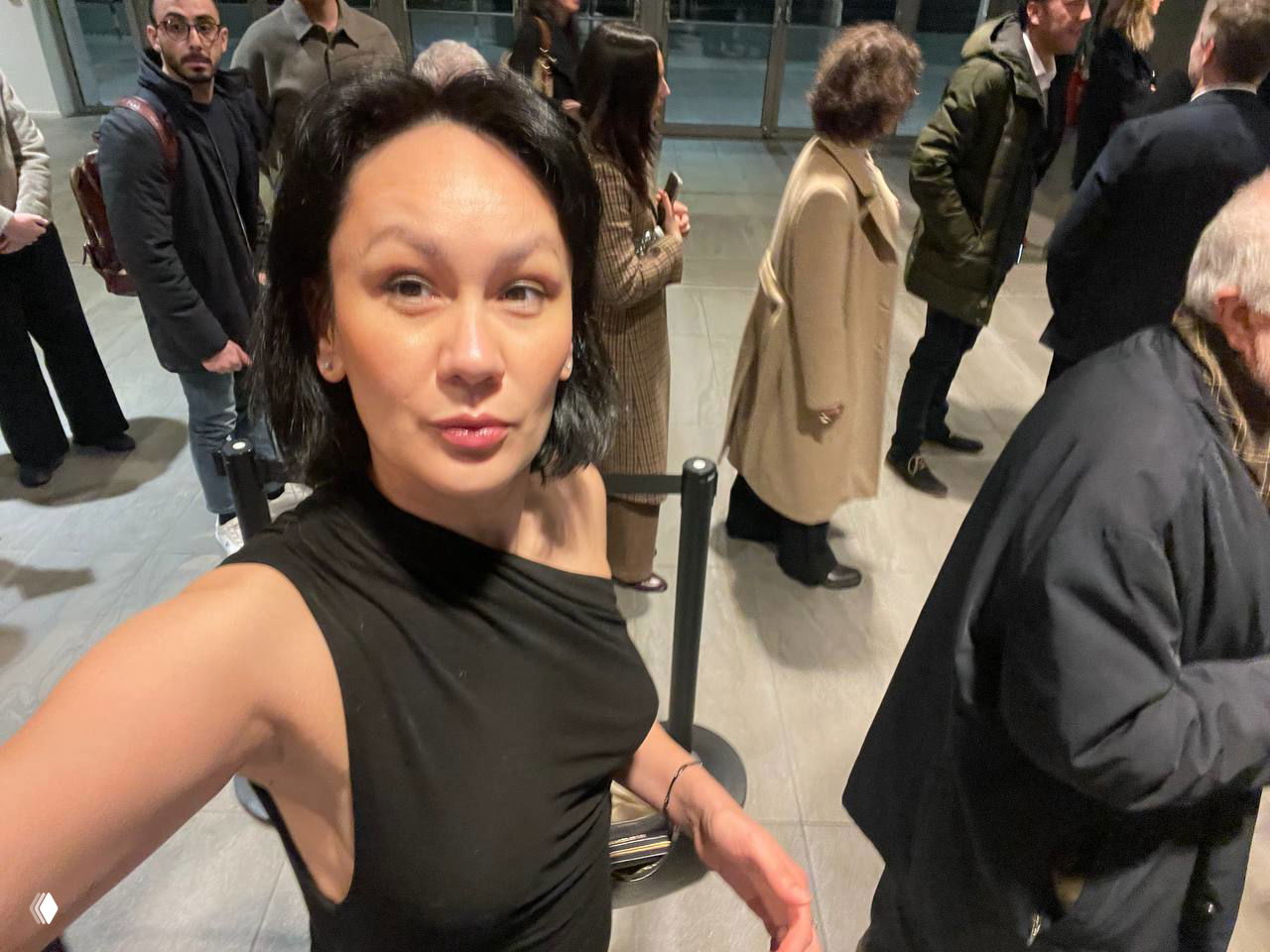 Селфи автора в фойе театра на фоне очереди и публики — чёрное платье, сумка через плечо, оживлённая атмосфера перед представлением.