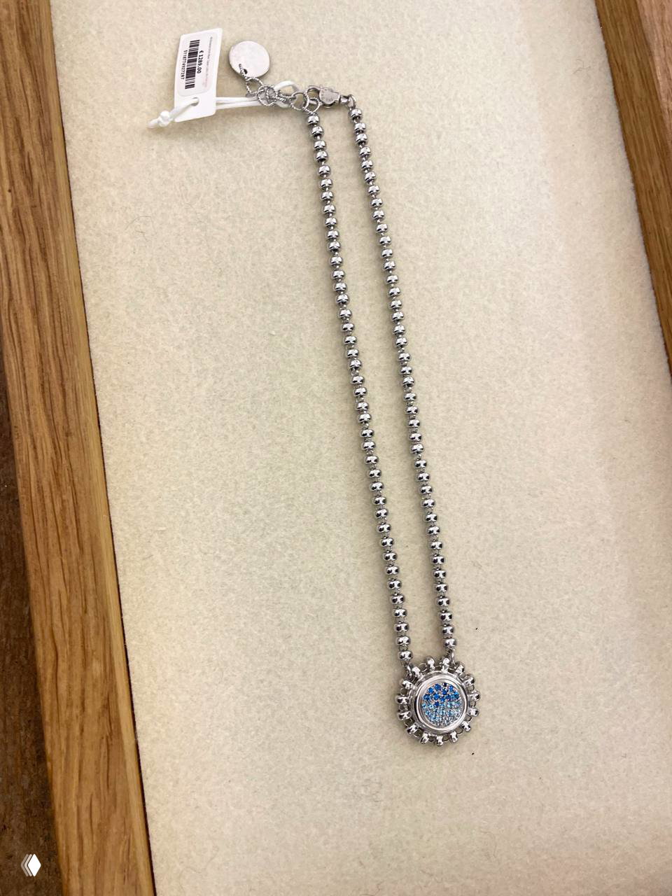 Крупный план тонкой серебристой цепочки с круглой подвеской и биркой — украшение на светлом подкладочном фоне в ювелирной витрине.