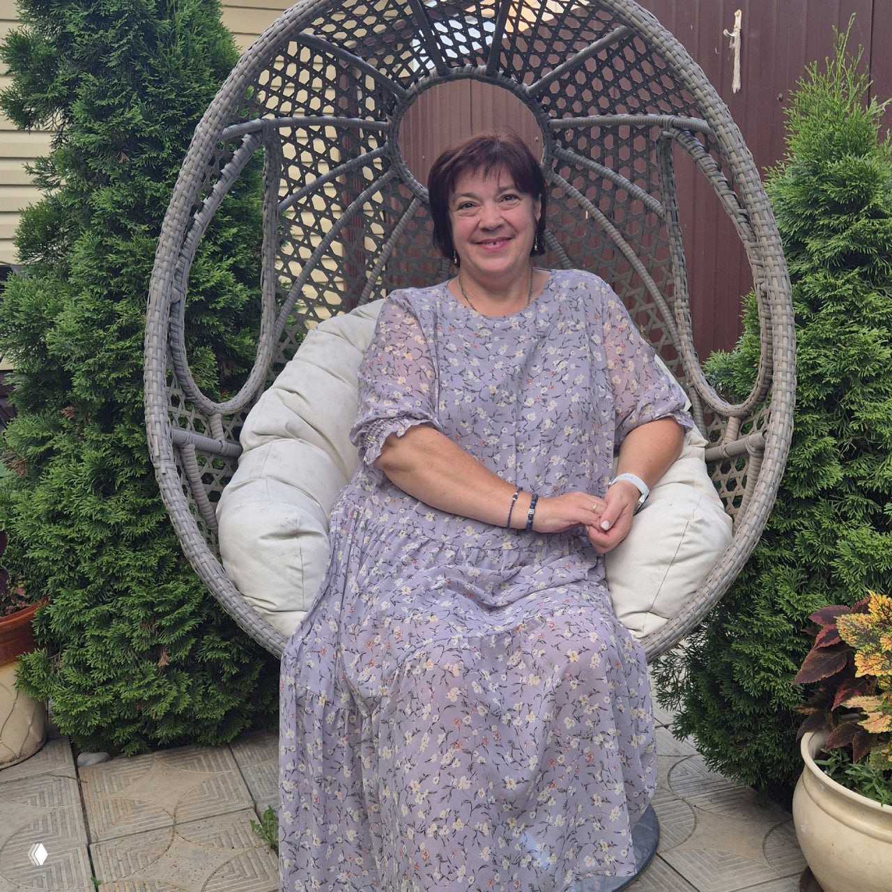 Женщина средних лет в лёгком платье сидит в плетёном подвесном кресле на фоне туй и забора, улыбается в садовой обстановке.