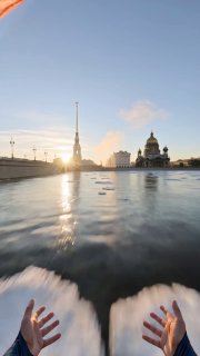 FPV-ракурс: руки внизу кадра над весенней Невой на рассвете, с плавно отражающимися льдинками, шпилем Петропавловской крепости и куполом Исаакия.