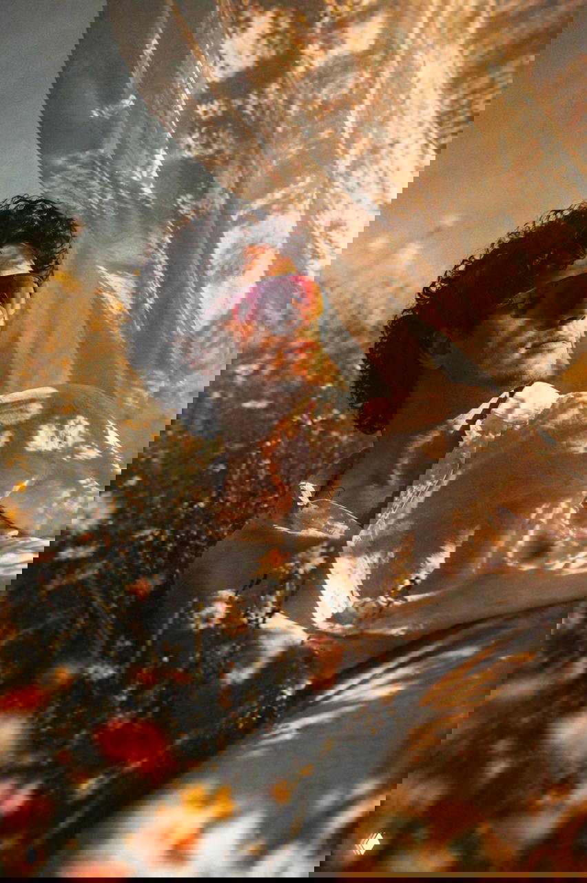 Портрет мужчины в белой рубашке и зеркальных очках рядом с развевающейся полупрозрачной вуалью на фоне летнего поля с цветами; мягкий золотой свет.