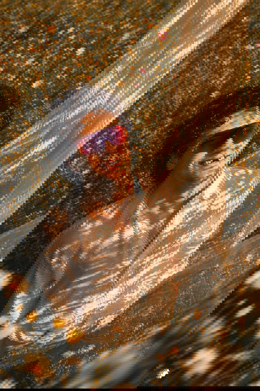 Мужчина в белой рубашке среди высоких полевых цветов, солнечные блики и тени от вуали создают текстуру на лице и одежде; ощущение присутствия и мечтательности.