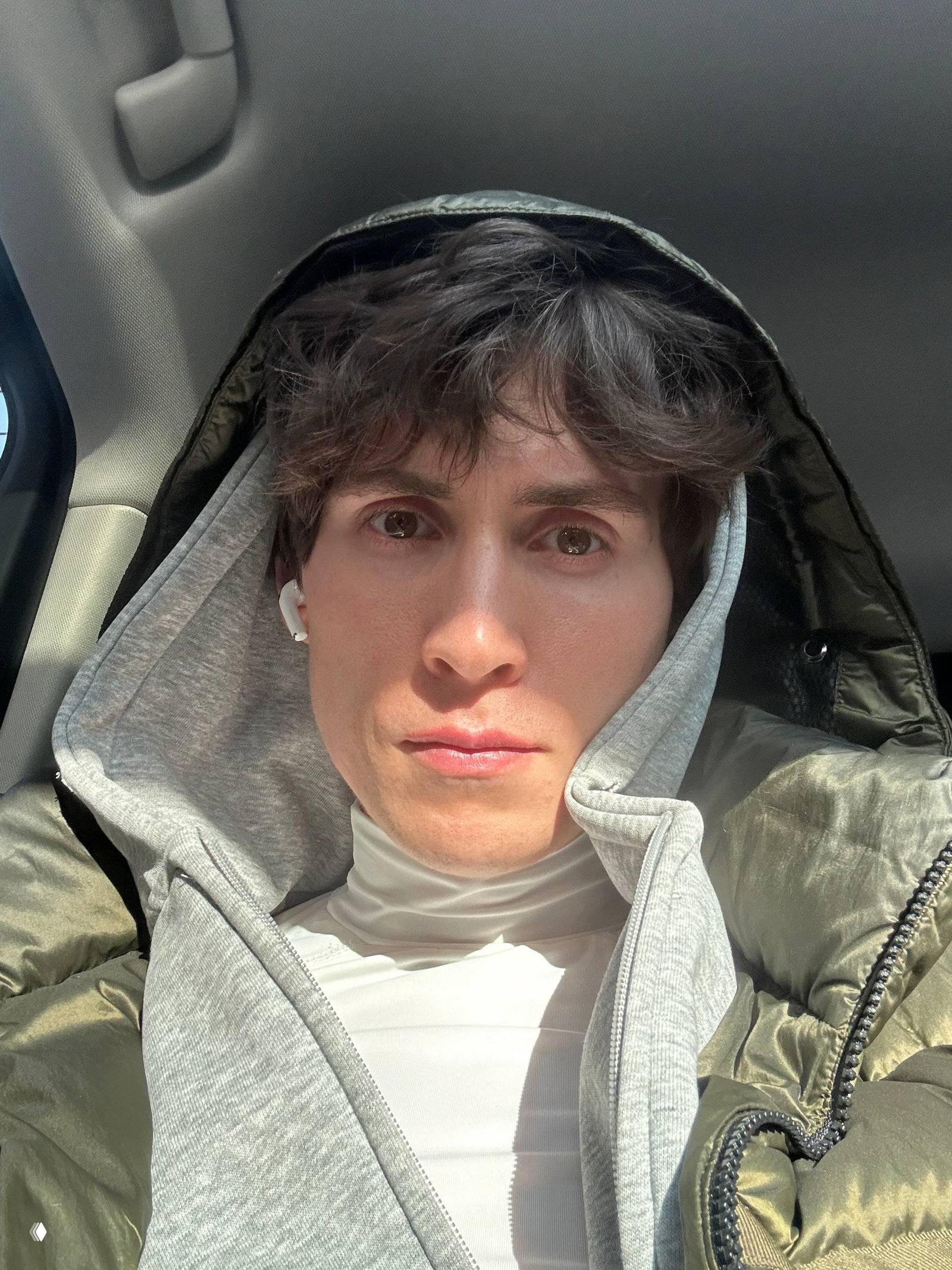 Селфи автора в капюшоне в салоне автомобиля при дневном свете, серьёзное выражение лица, один наушник AirPod виден в ухе.