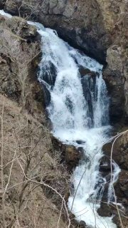 Водопад в скалистом овраге: талая вода стремительно сбегает между обнажёнными весенними кустами и камнями.