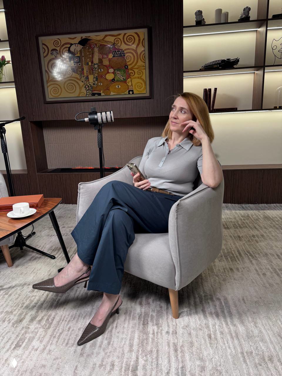 Автор канала задумчиво сидит в кресле с телефоном в руках рядом с микрофоном; на столике видна чашка, виден интерьер и реквизит студии.