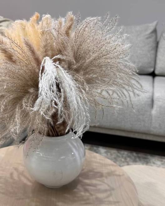 Ваза с высокой сухой травой (пампасной) стоит на столе на фоне серого дивана; декоративный элемент, создающий мягкую атмосферу интерьера.