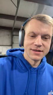 Селфи мужчины в синей толстовке и наушниках в мастерской: крупный план лица на фоне металлической конструкции ангара.