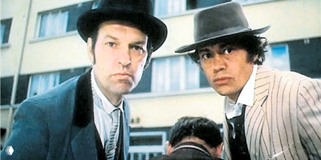 Кадр из старого фильма: двое мужчин в шляпах и плащах на фоне жилого дома, атмосферный ретро-образ с нюансами ретро-эстетики