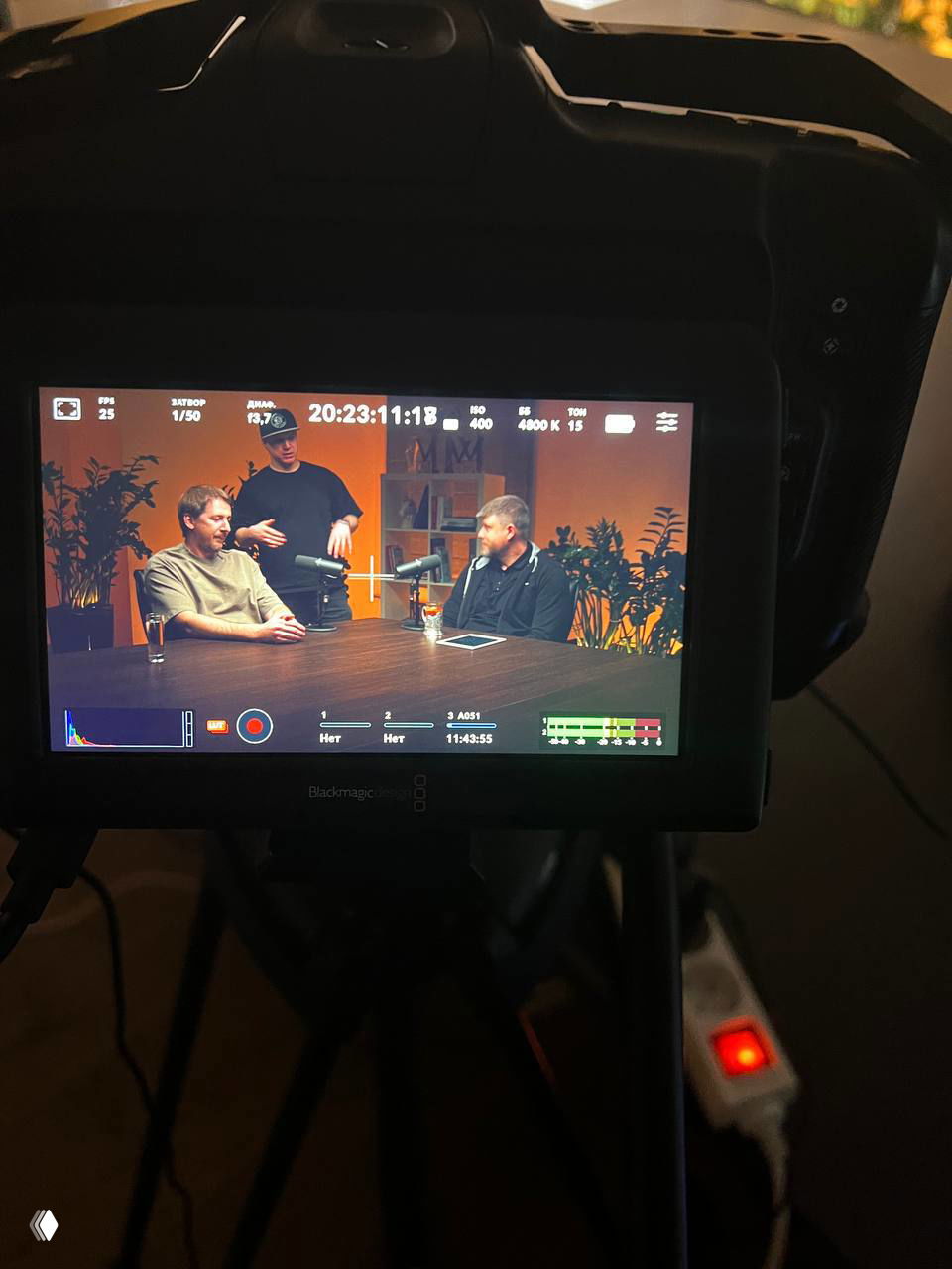 Экран камеры показывает кадр с двумя участниками подкаста за столом, на мониторе видна композиция сцены и элементы интерфейса записи.