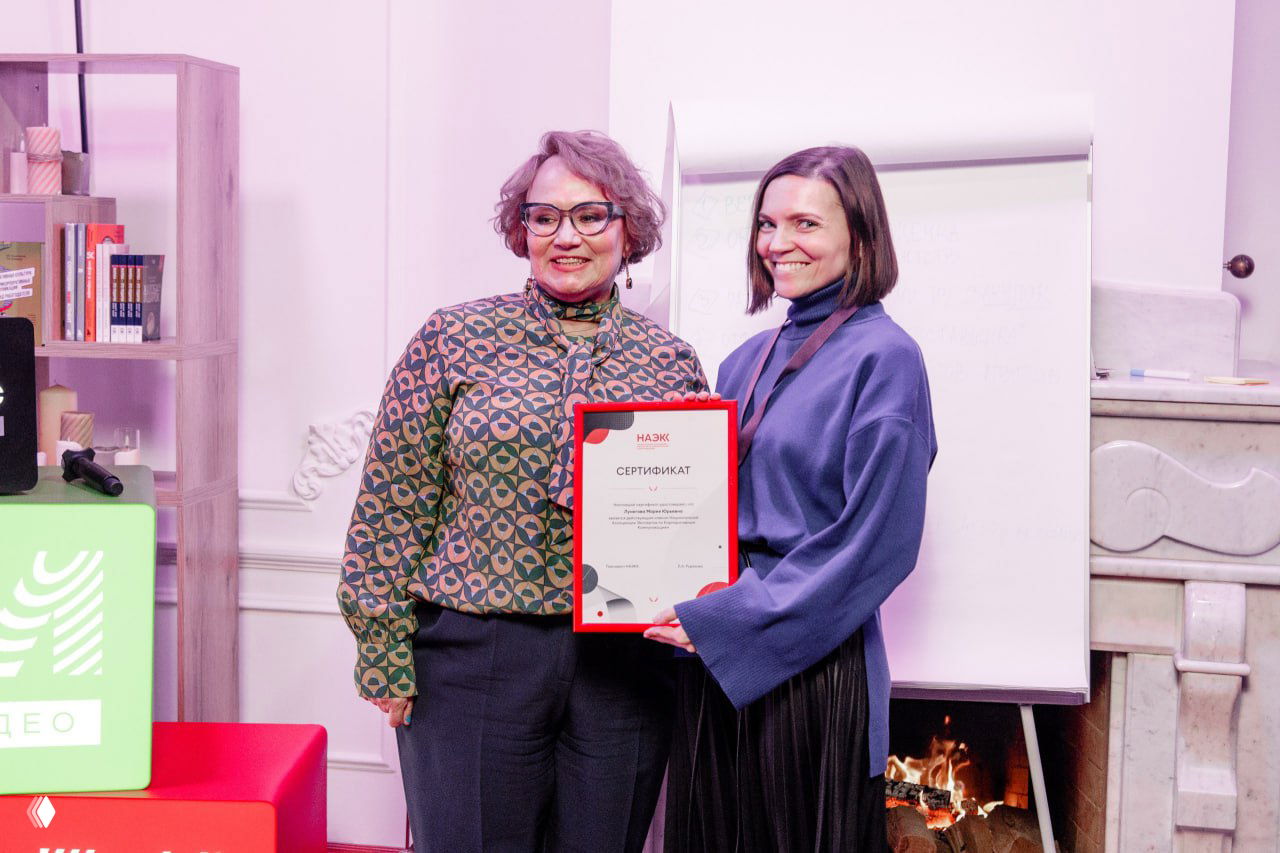 Две участницы принимают сертификат на встрече НАЭКК: вручение диплома на фоне камина, флипчарта и интерьерной отделки