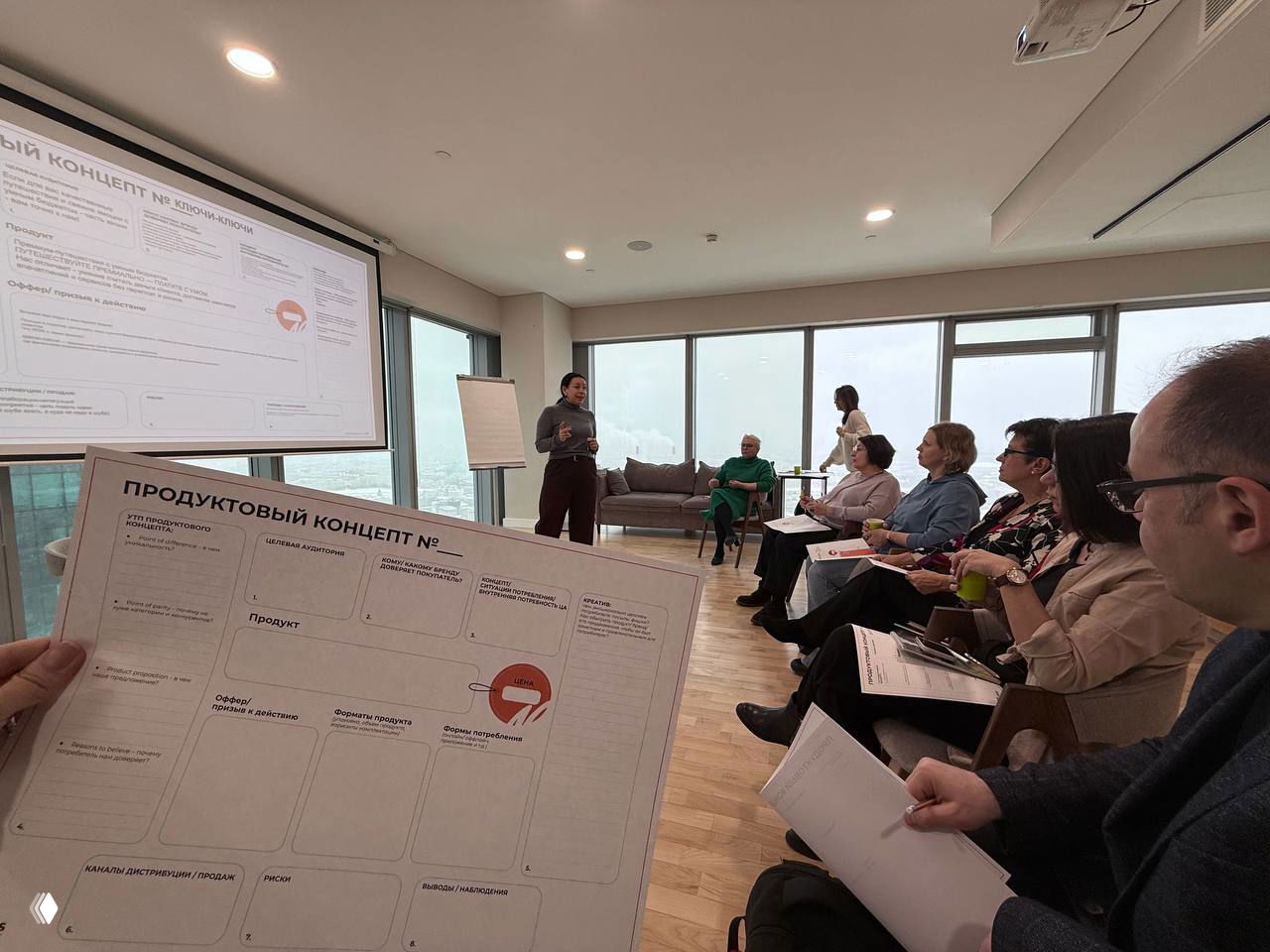 Вид из рук участника на шаблон «Продуктовый концепт» во время сессии: лист с блоками на фоне аудитории и большого проектора в комнате с окнами.