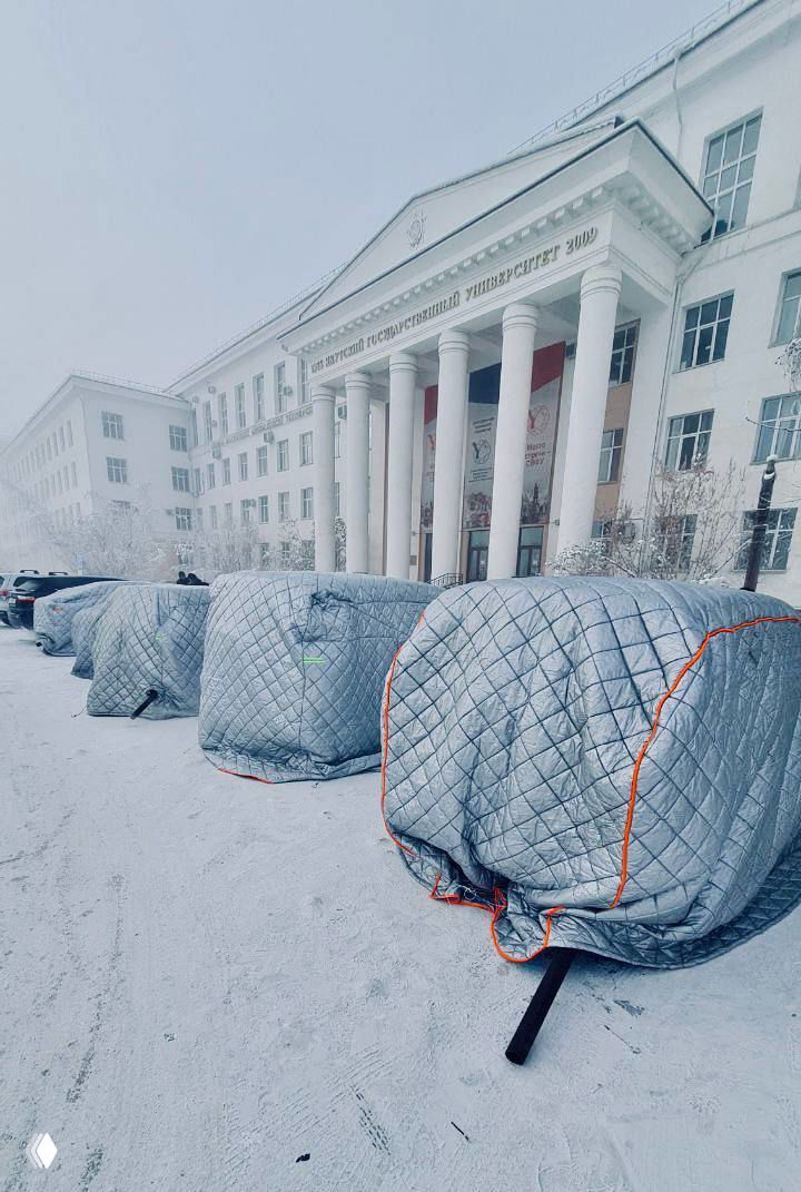 Ряд автомобилей на парковке в Якутске, укрытые стёгаными чехлами и покрывалами, вид на здание университета на заднем плане, техника защиты от мороза.