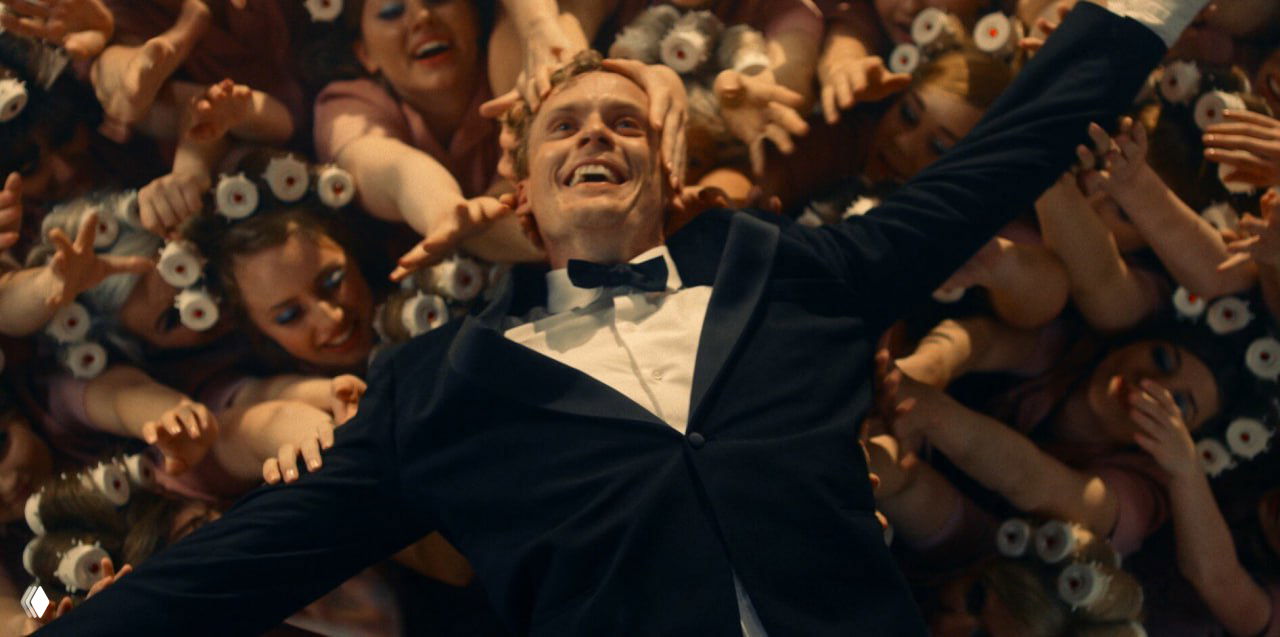 Кадр из сериала: мужчина в смокинге лежит среди множества рук и бигуди, выражение восторга и театральная композиция сцены.