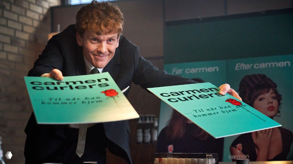 Сцена с рекламными плакатами «carmen curlers»: мужчина в костюме раздаёт листовки на фоне постеров с изображениями женщин.