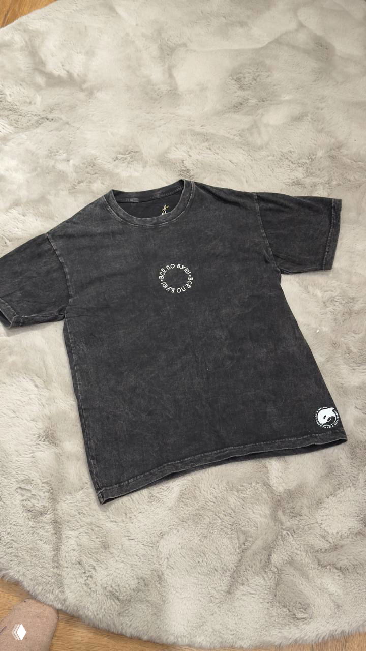 Черная футболка с эффектом «стирки» и круглым белым логотипом на груди, разложена на сером пушистом фоне — фото модели футболки.