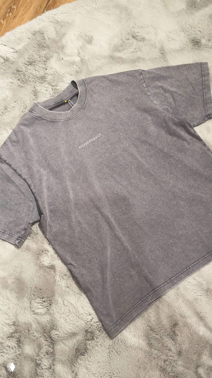 Темно‑серая футболка с едва заметным белым логотипом в центре, разложена на пушистом ковре — фото варианта коллекции.