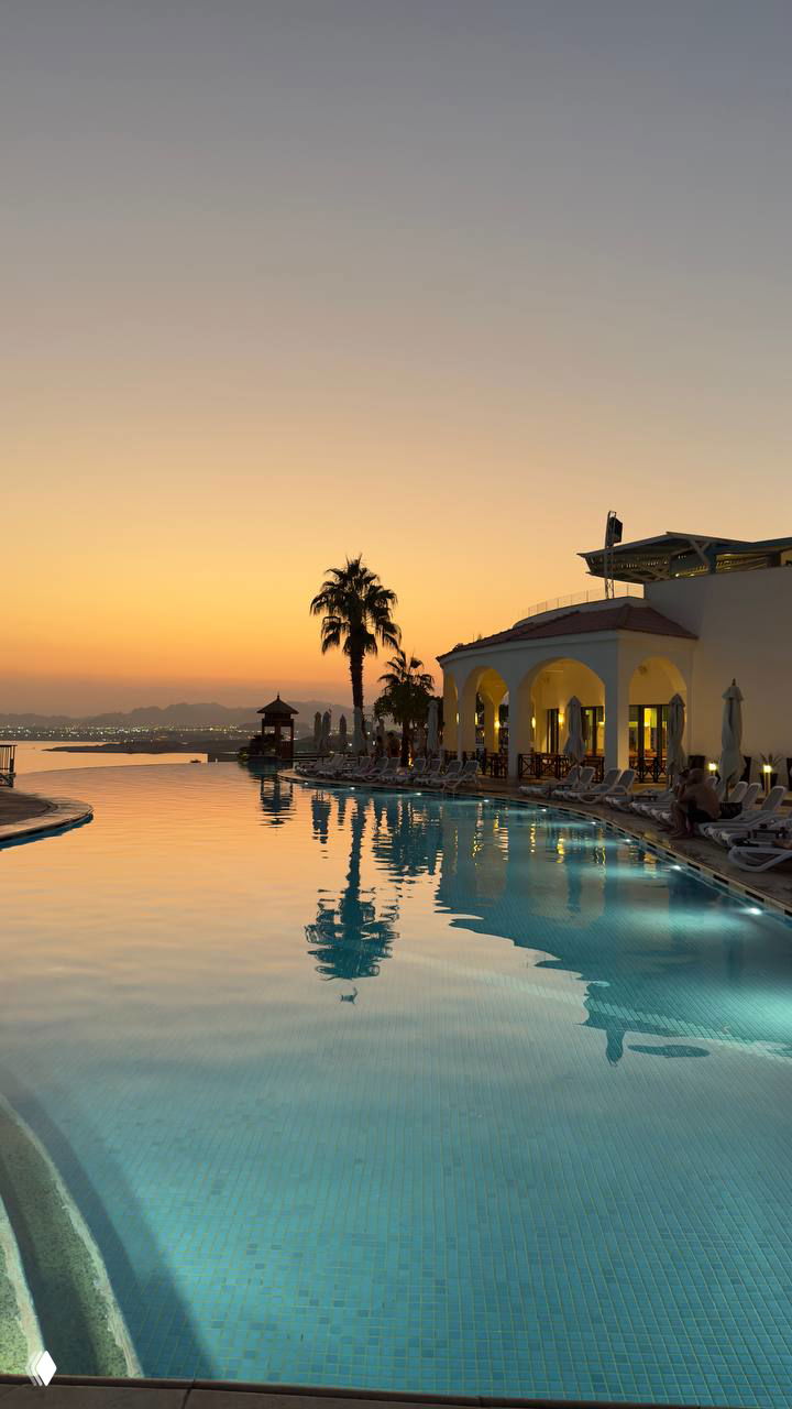 Бассейн и береговой отель в Шарме на закате с отражением пальмы и здания в спокойной воде; тёплые оранжево-лиловые тона неба.