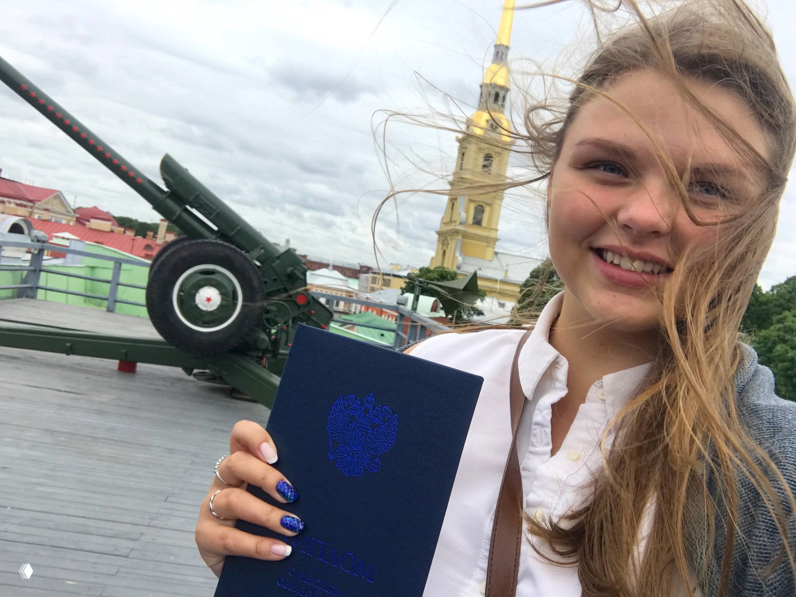 Селфи с синим дипломом у Петропавловской крепости: девушка держит диплом на фоне пушки и шпиля Петропавловского собора, ветер в волосах.