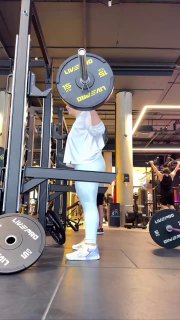 Спортзал: та же женщина выполняет присед со штангой, ракурс ближе к фронту; видно вынос коленей, стойку и расстановку ног во время сета.