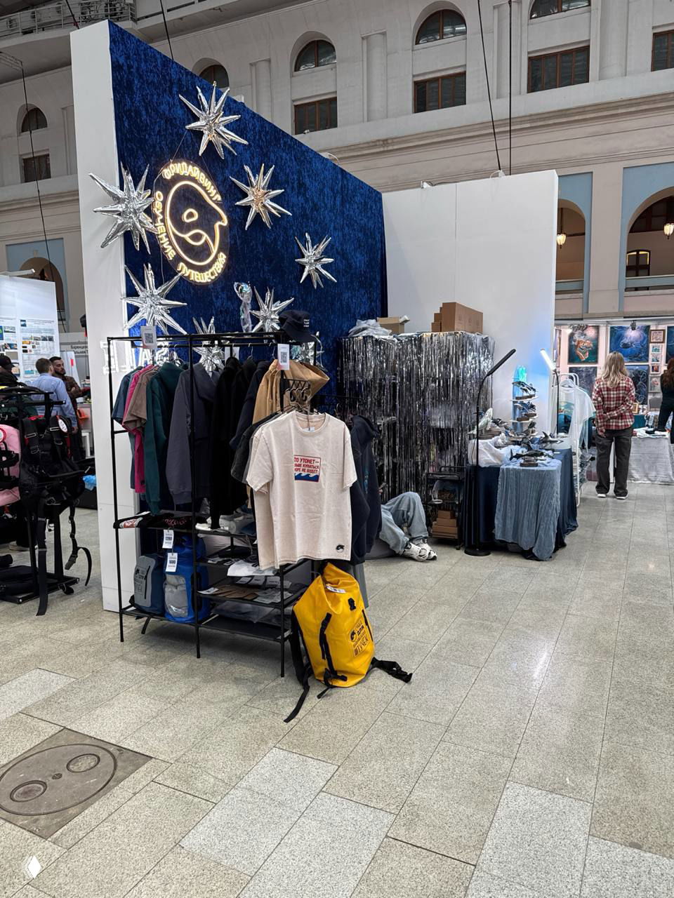 Широкий кадр стенда: стойка с футболками и куртками, витрина мерча и яркий жёлтый туристический рюкзак на полу в зоне Moscow Dive Show