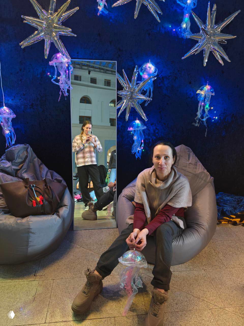 Уютная зона стенда: человек сидит в пуфе и держит декоративную медузу, на фоне зеркало с отражением автора и подвесные светящиеся звёзды‑медузы