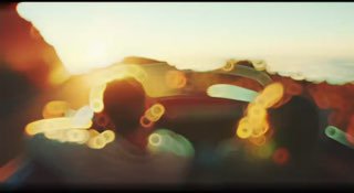 Кадр из документалки: размытый силуэт человека за рулём автомобиля на фоне тёплого закатного света и ярких бликoв.