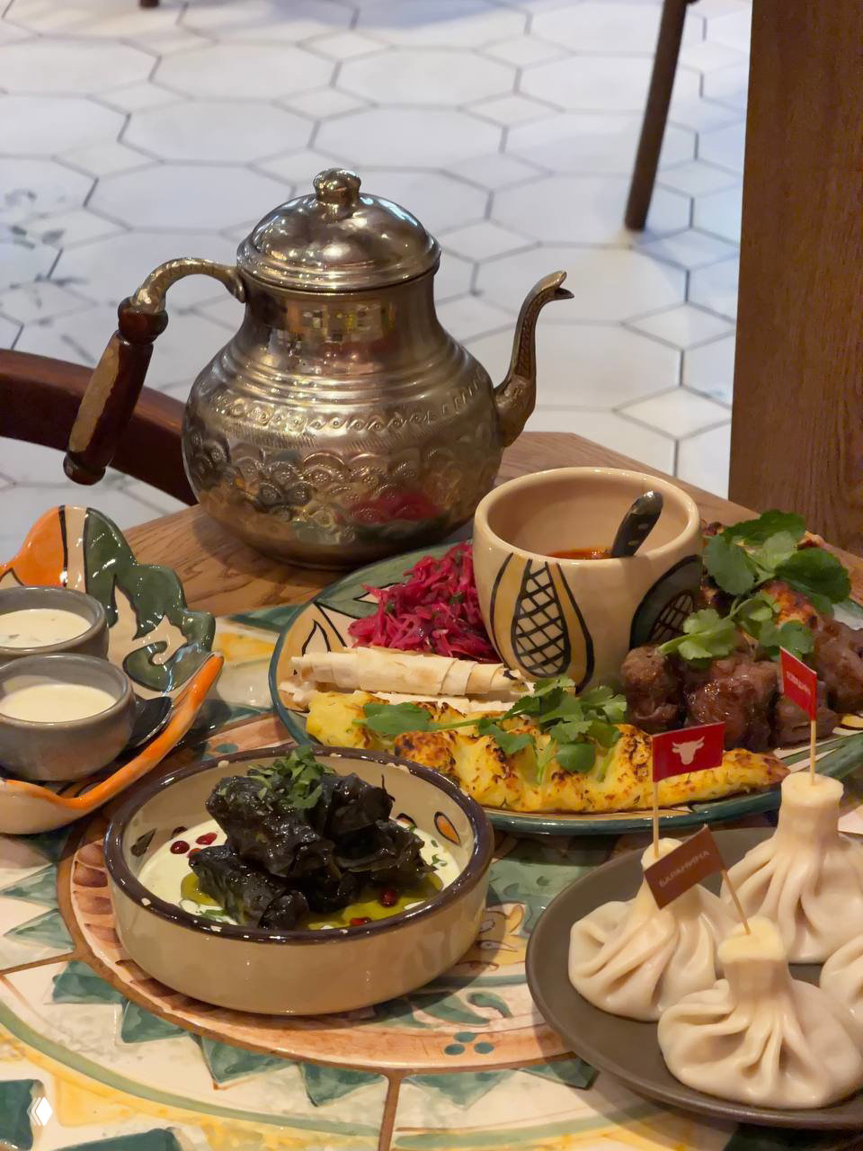 Подача основных блюд: хинкали, мангальные блюда и соусы на тарелках, рядом металлический чайник — праздничная сервировка грузинских блюд.