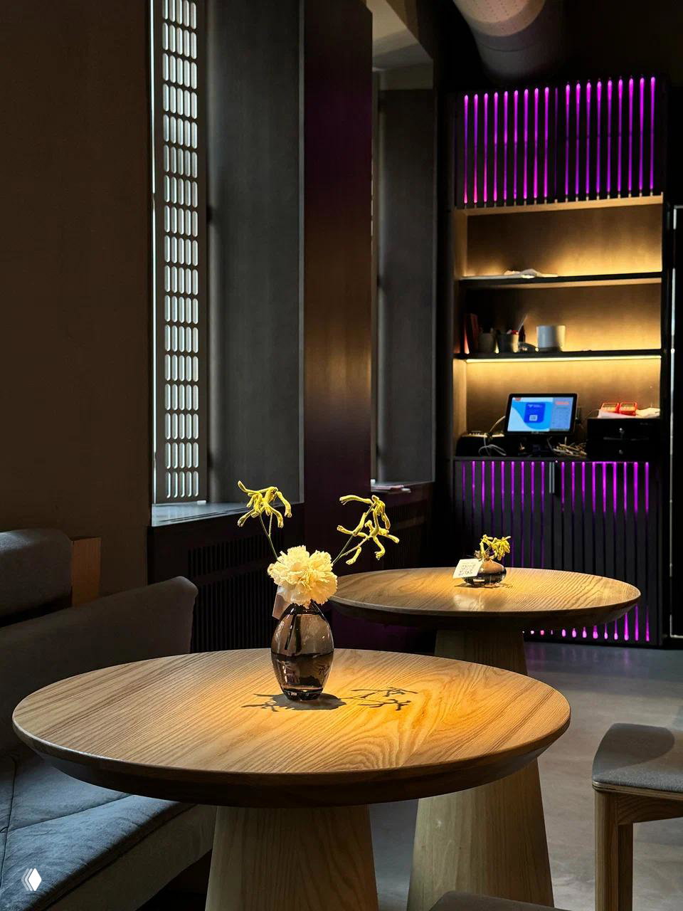 Интерьер бара Tsunami: деревянные столы с небольшими вазами, приглушённое освещение и фиолетовые декоративные рейки на заднем плане.